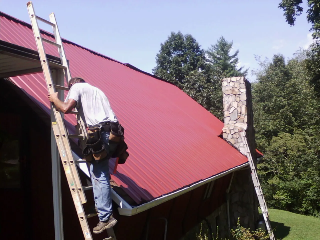 Roofer on a ladder beside a red metal roof and stone chimney — Soffit & Fascia Repair by Roofing Brothers in Eastanollee
